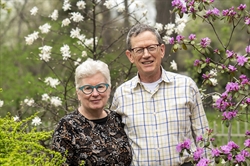 [ai] A woman with curly white hair and glasses stands next to a man wearing a plaid shirt in a garden. They are surrounded by blooming white and pink flowers, creating a vibrant natural backdrop.